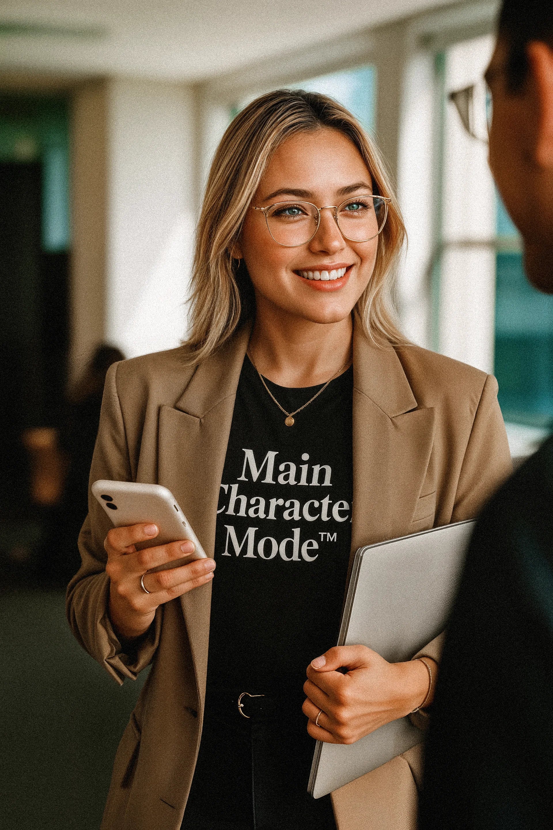Woman in a professional setting holding a phone and a laptop, wearing a shirt with text.